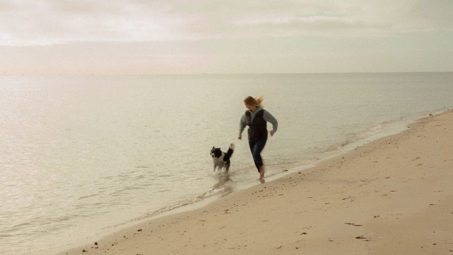 Woman running along a calm sandy beach with a black and white border collie dog at the shoreline.