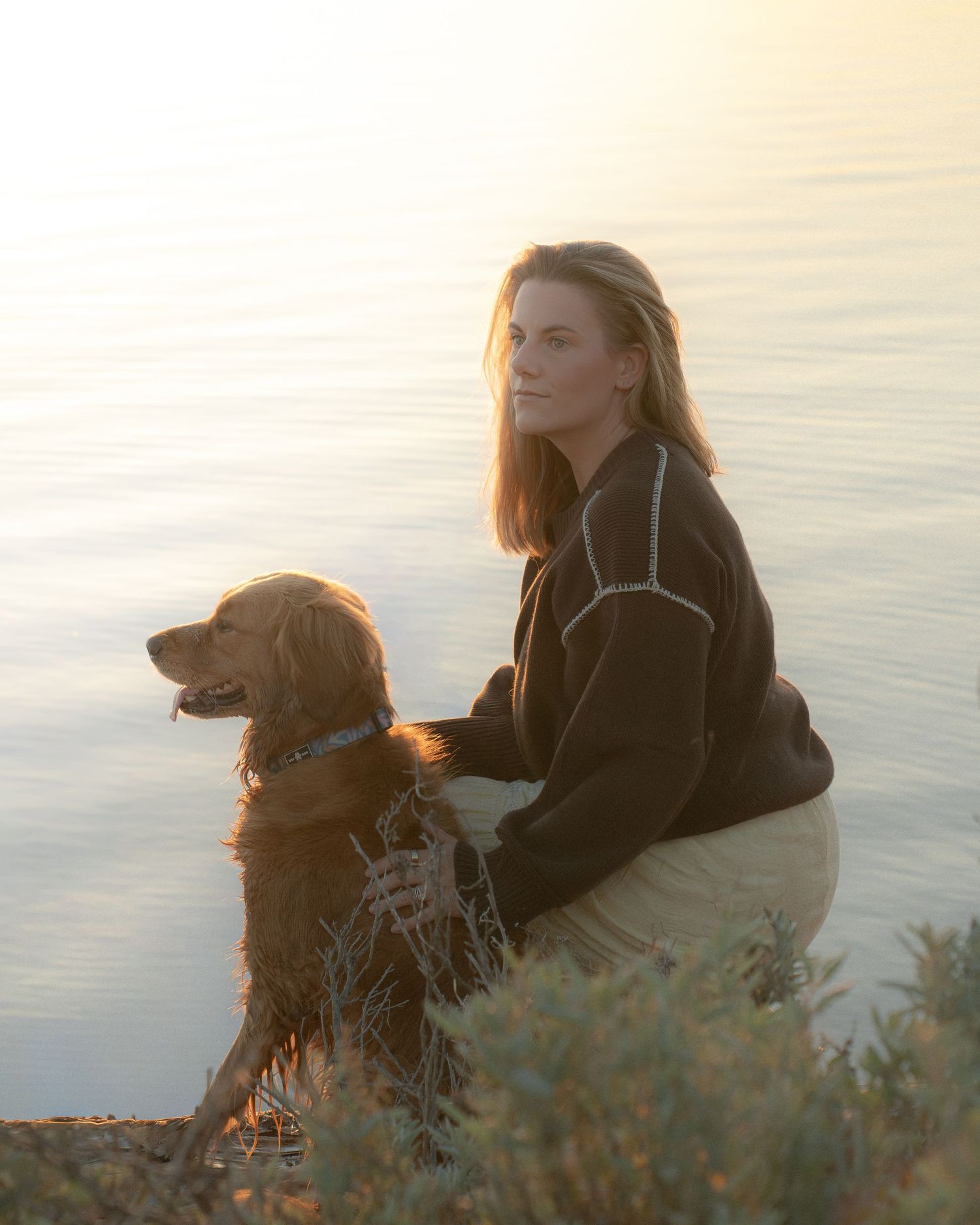 Woman sitting with her golden retriever dog by a glowing lake at sunset.