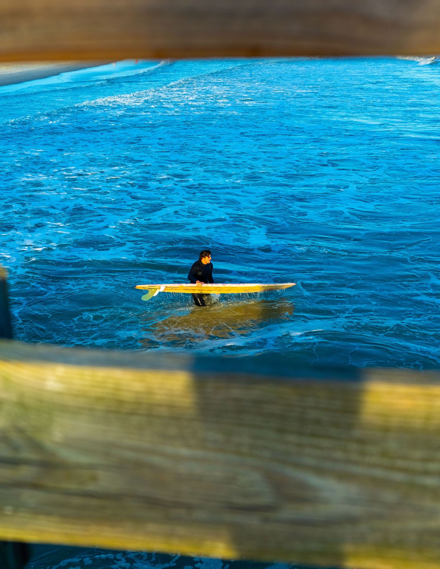 Action sports photography of a surfer on a yellow board in deep blue ocean water, captured through a wooden pier frame by Shane Anthony.