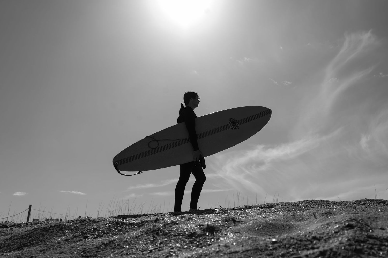 Moody black and white silhouette of a surfer carrying a longboard on a cliffside, showcasing cinematic documentary style by Shane Anthony.