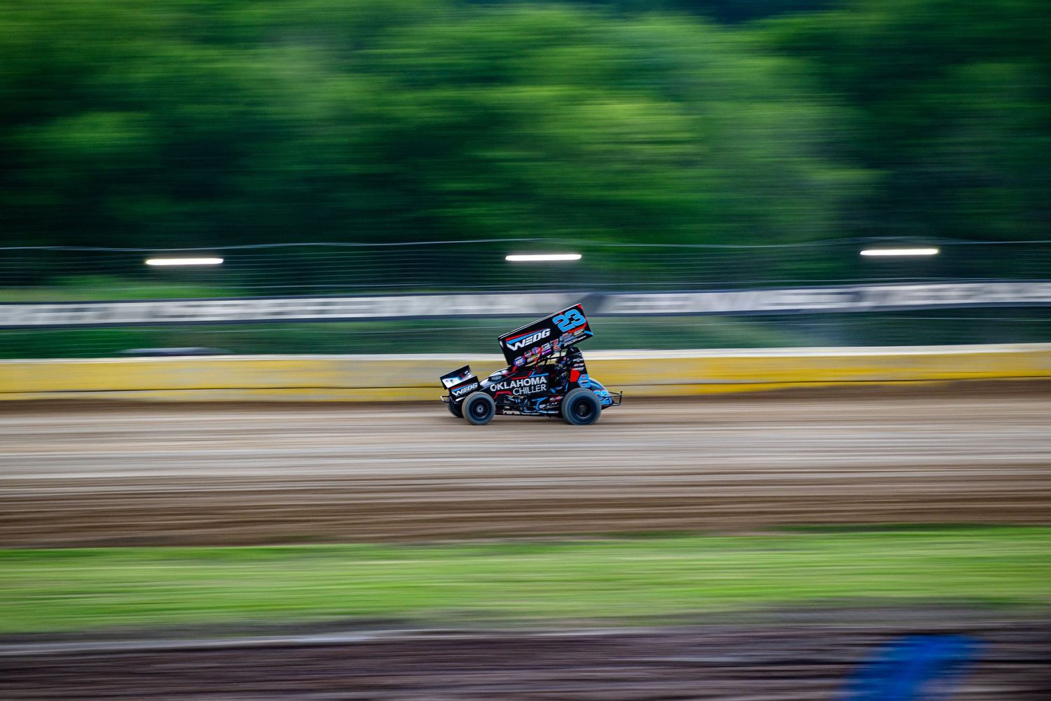 Sprint car racing on a dirt track with vibrant green trees in the background.