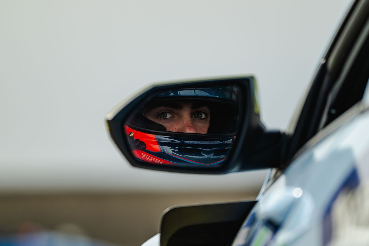 A racing driver wearing a helmet reflected in a car side mirror on a motorsport track.