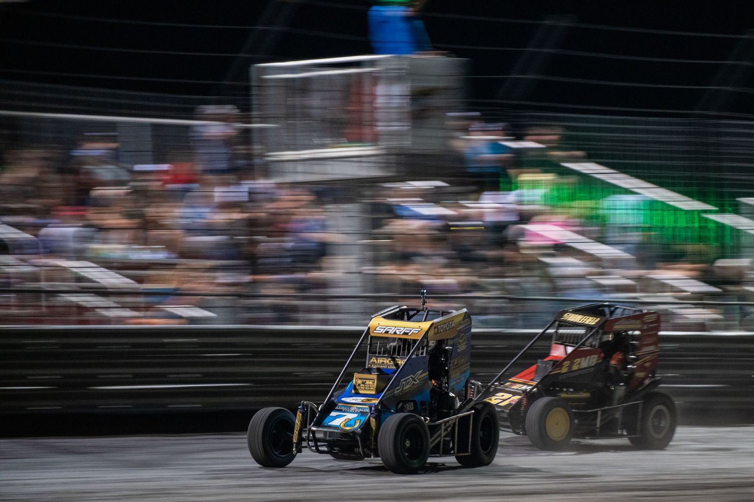 Two midget race cars competing side by side on an indoor dirt track at night.