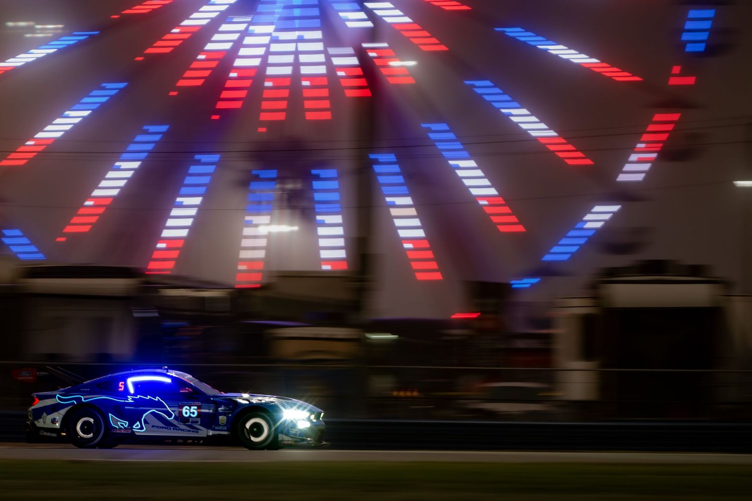 Blue racing car speeds past colorful starburst light display at night.