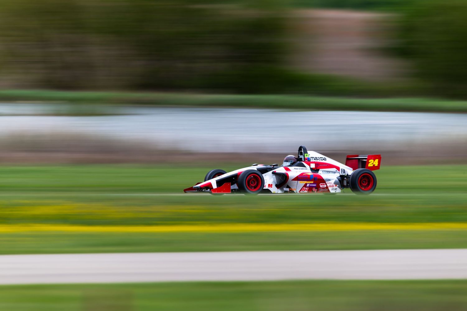 Red and white Formula Mazda number 24 racing at high speed on a road course.