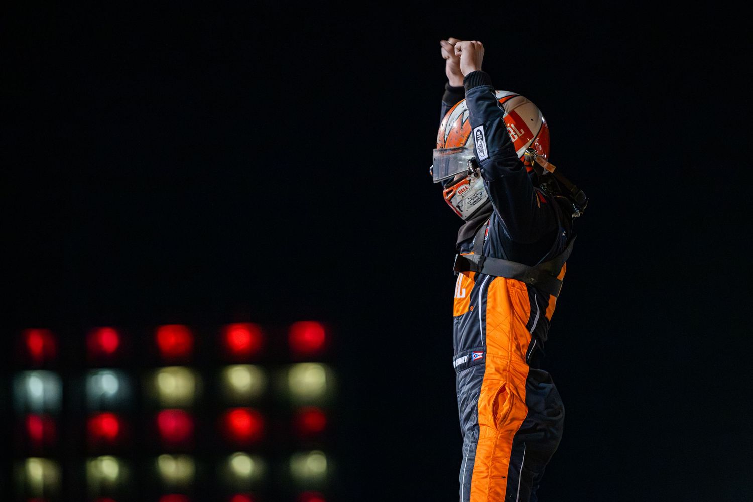 Racing driver in orange suit standing on car celebrating victory under night lights.