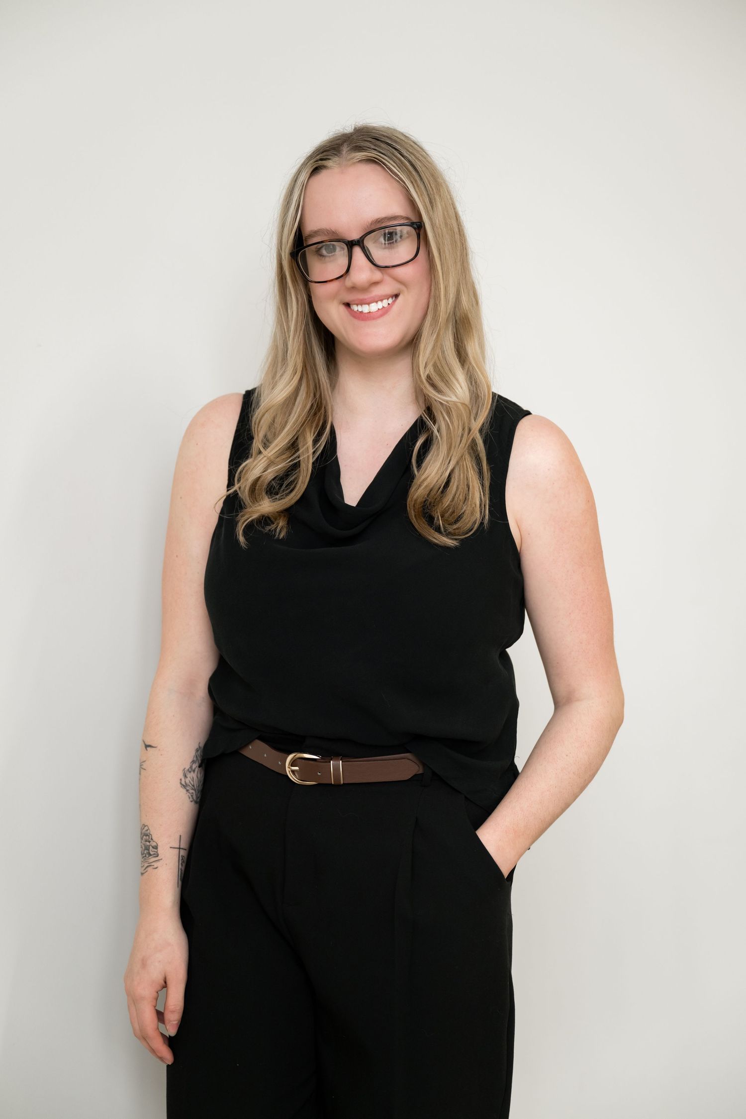 Smiling blonde woman with glasses wearing black sleeveless top and pants against white background.