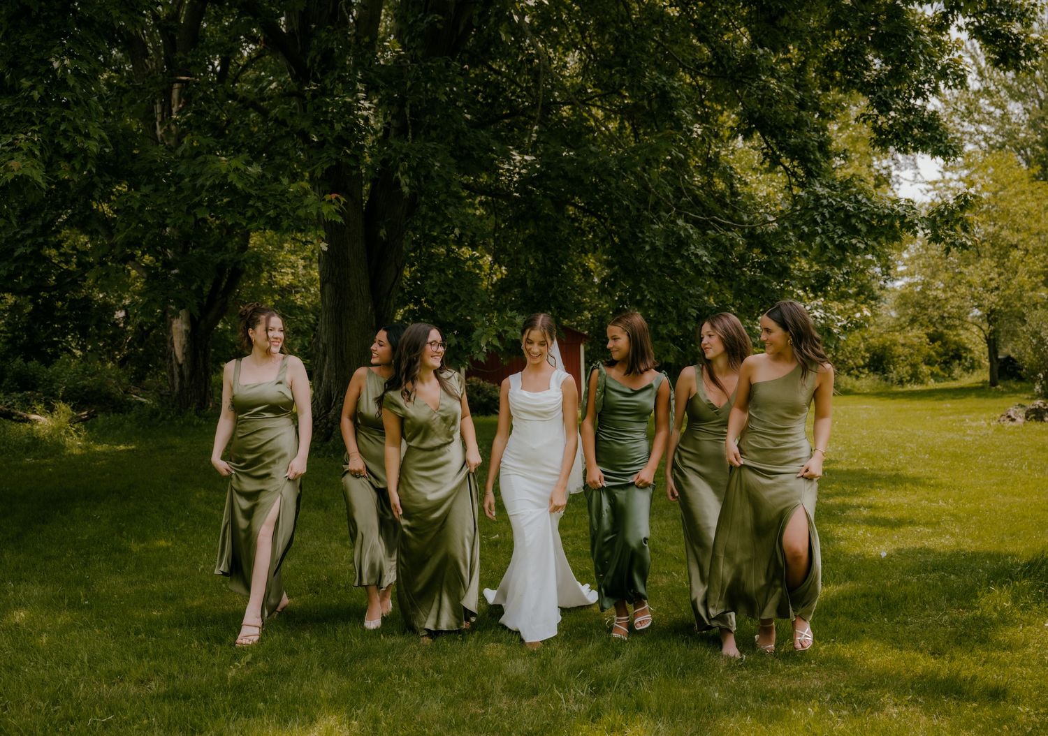 Bride in white gown surrounded by six bridesmaids in sage green satin dresses under large trees.
