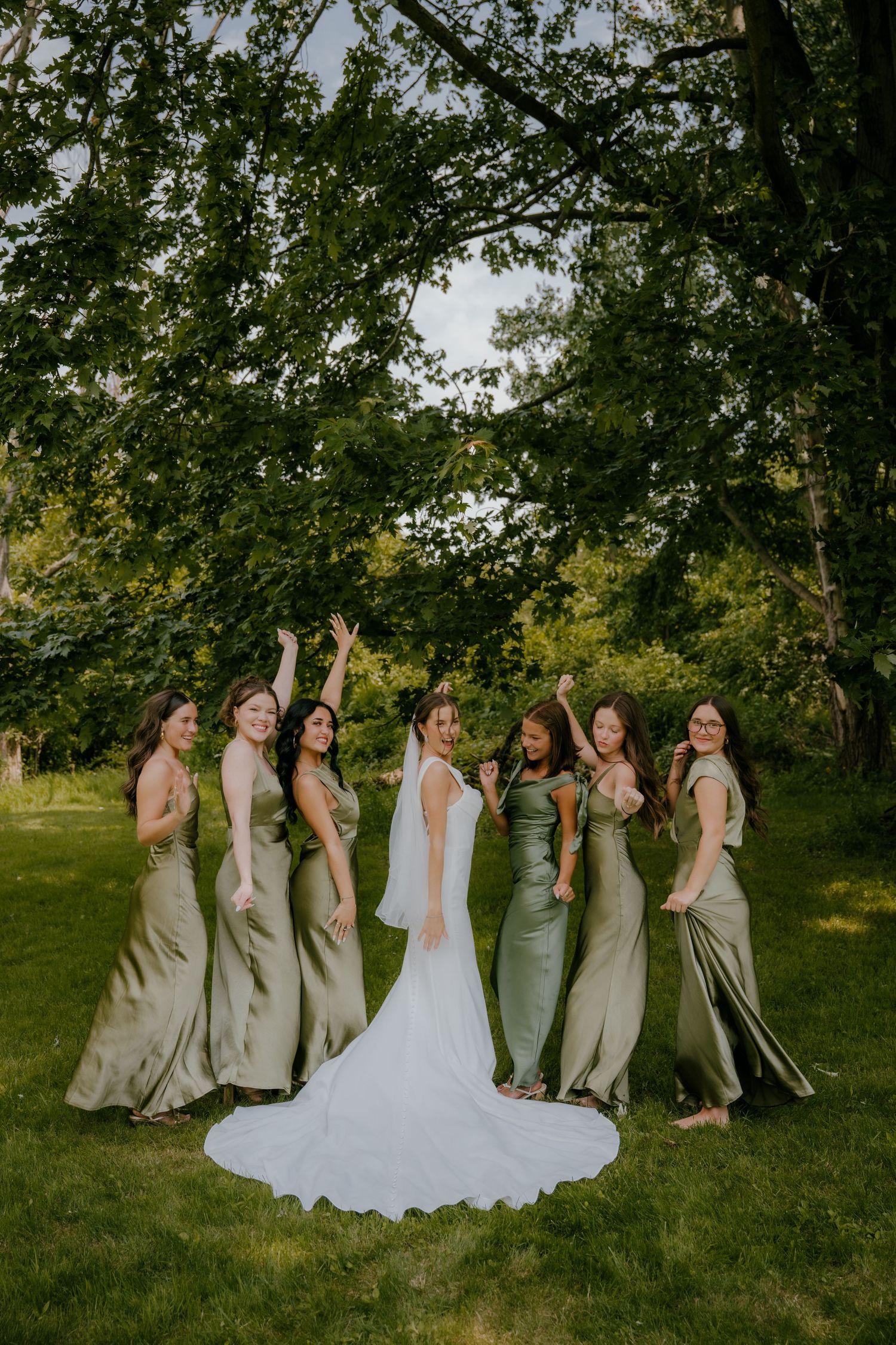 Bride with bridesmaids in sage green satin gowns celebrating outdoors under lush green trees.