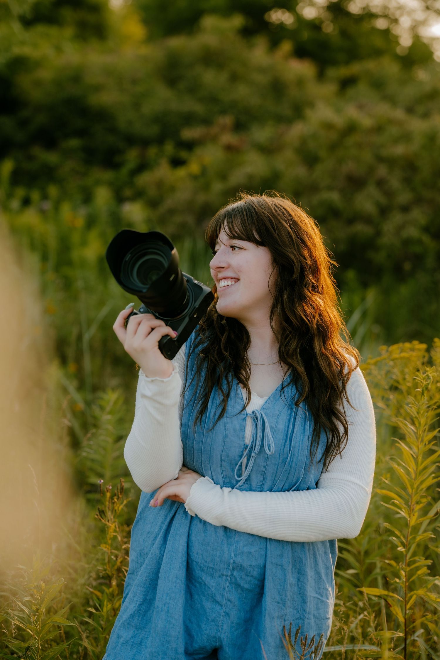 Smiling female photographer holding a DSLR camera outdoors in a golden hour field.