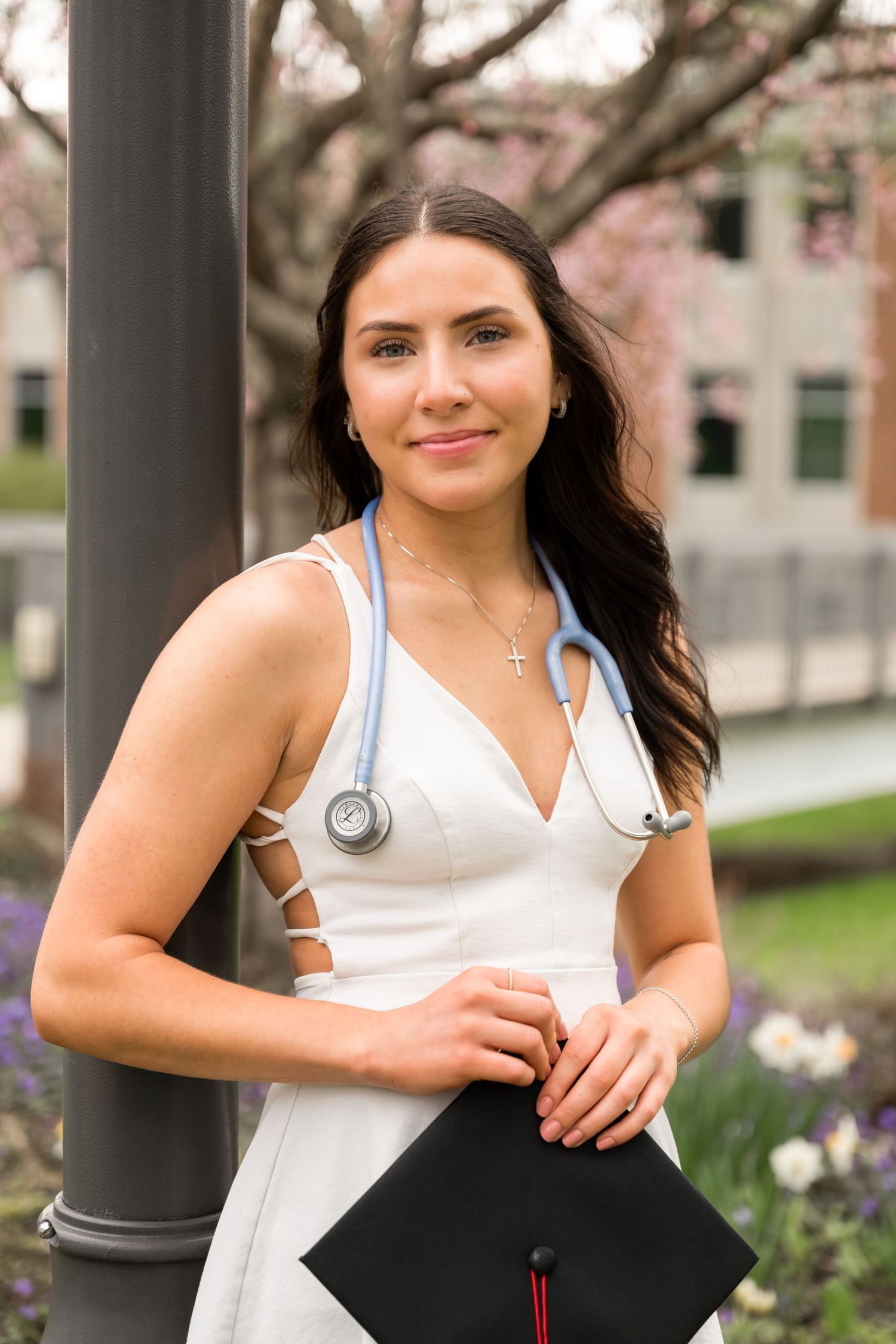 Female nursing graduate in white dress with stethoscope holding graduation cap outdoors.