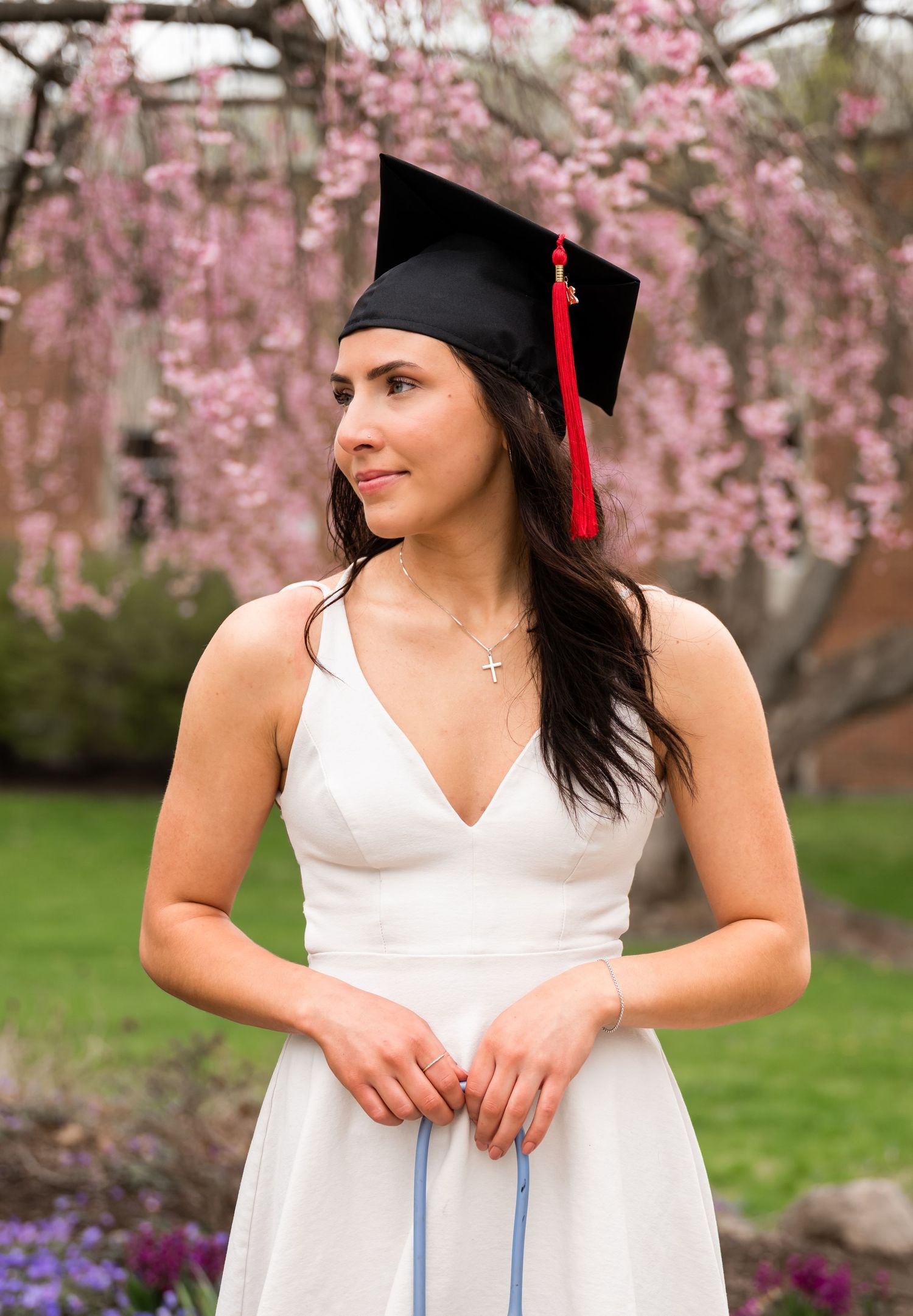 Young woman in white dress and black graduation cap with red tassel posing near blooming cherry trees.