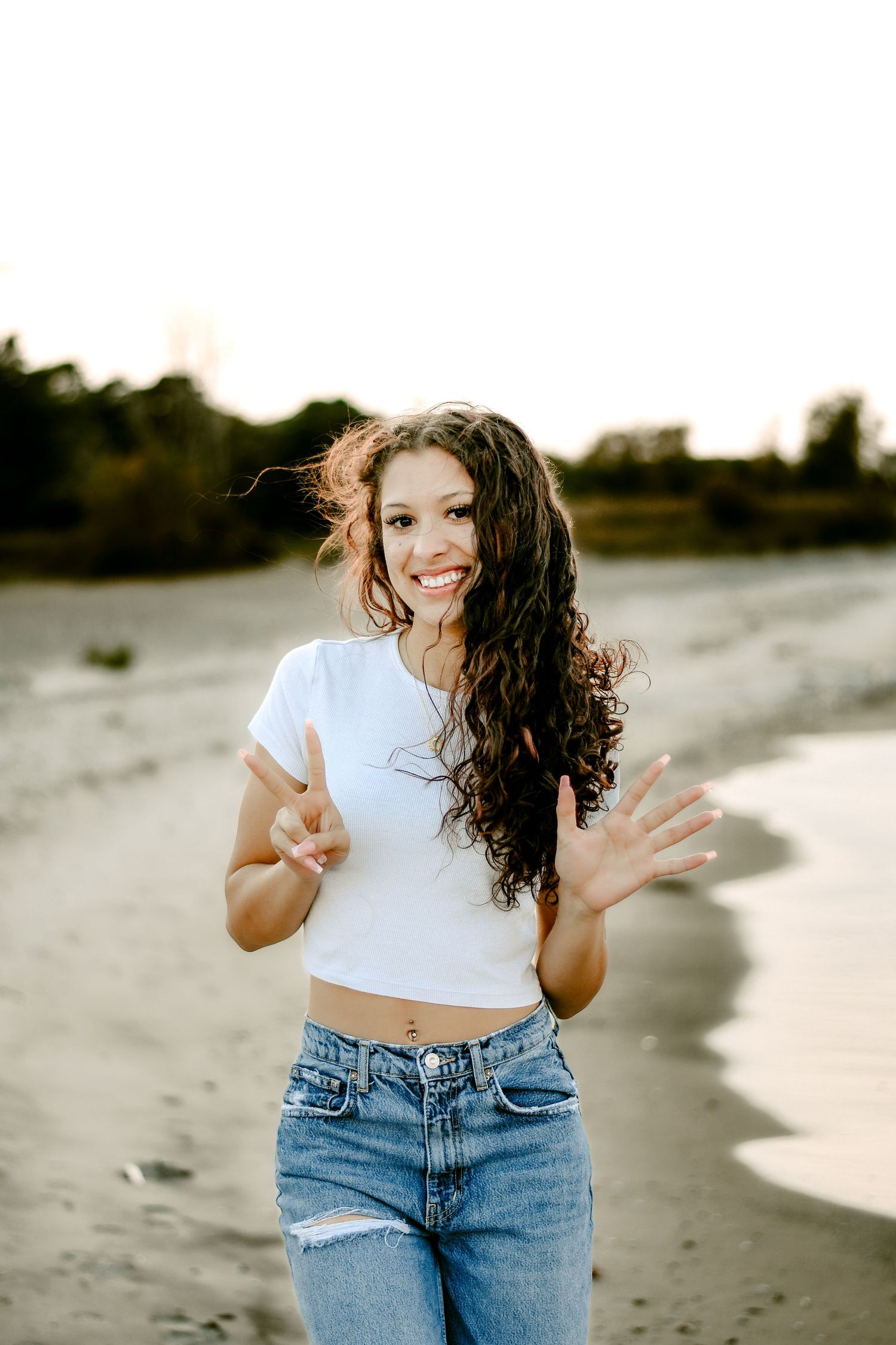 Smiling young woman with curly hair making peace sign on a sandy beach at sunset.