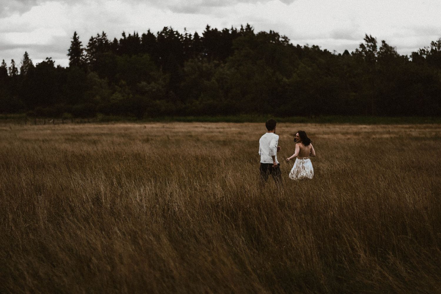 Couple walking hand in hand through golden tall grass field with dark forest backdrop.