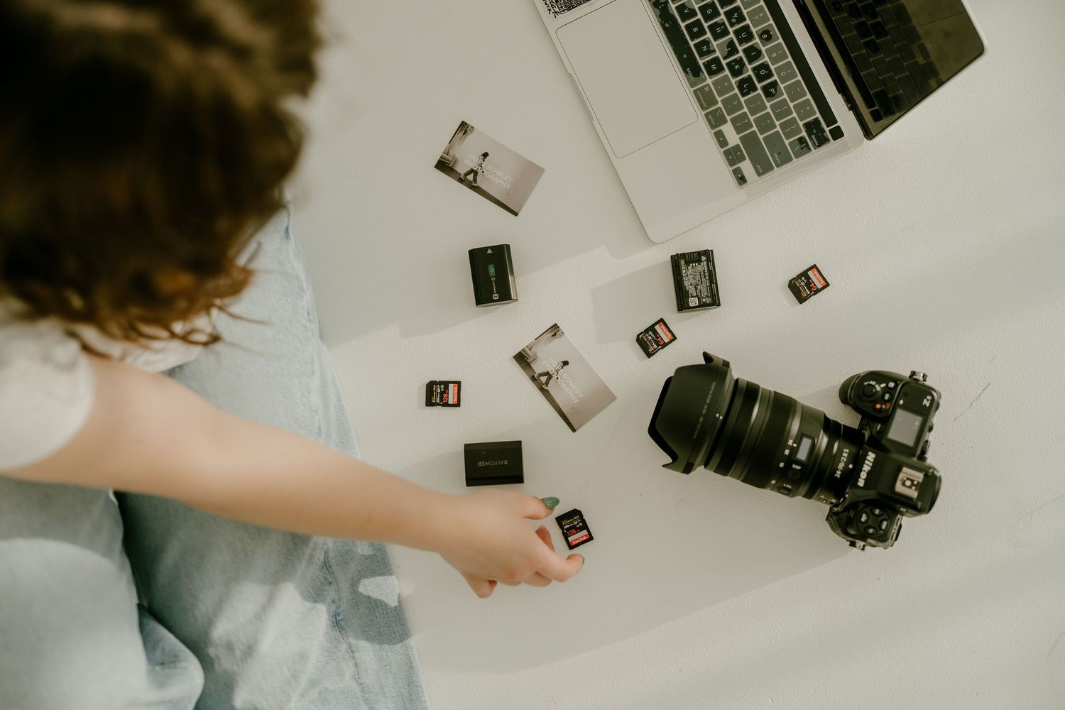 Photographer organizing SD memory cards beside a DSLR camera and laptop on a white desk.