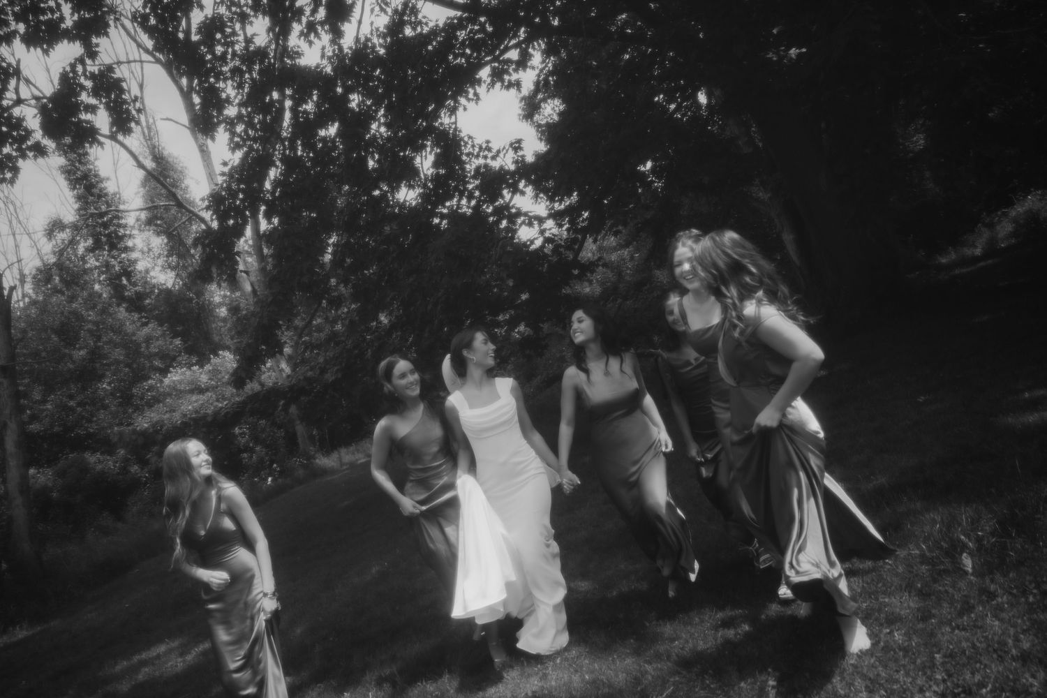 Black and white candid photo of bride and bridesmaids laughing and walking through a wooded area.