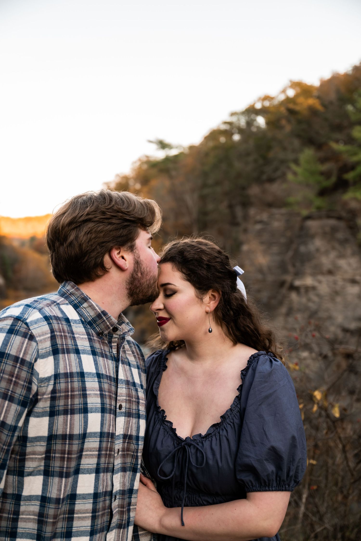 Couple sharing a tender forehead kiss during golden hour engagement session on rocky hillside.