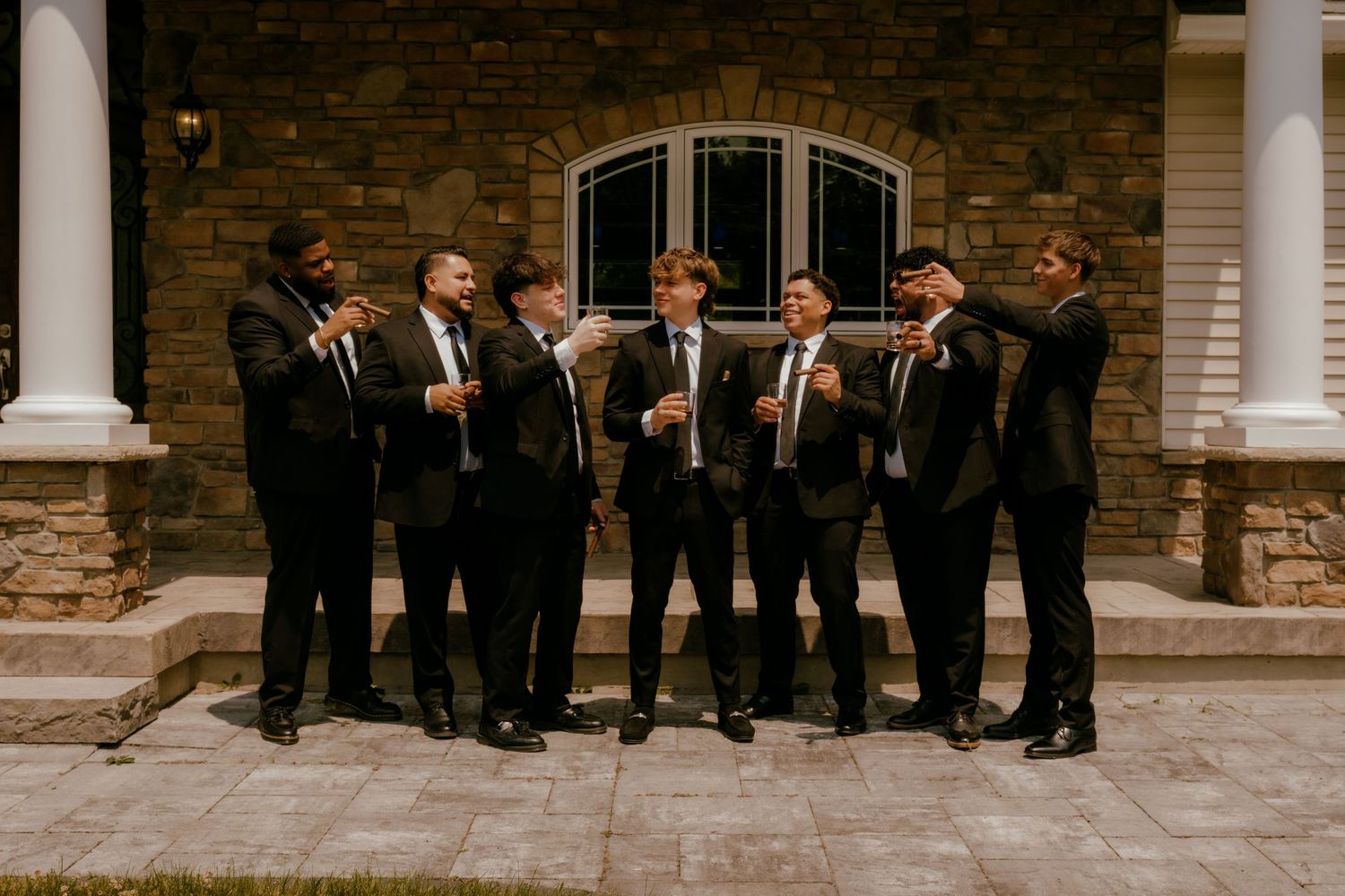 Group of groomsmen in black suits laughing and celebrating outside a stone building.