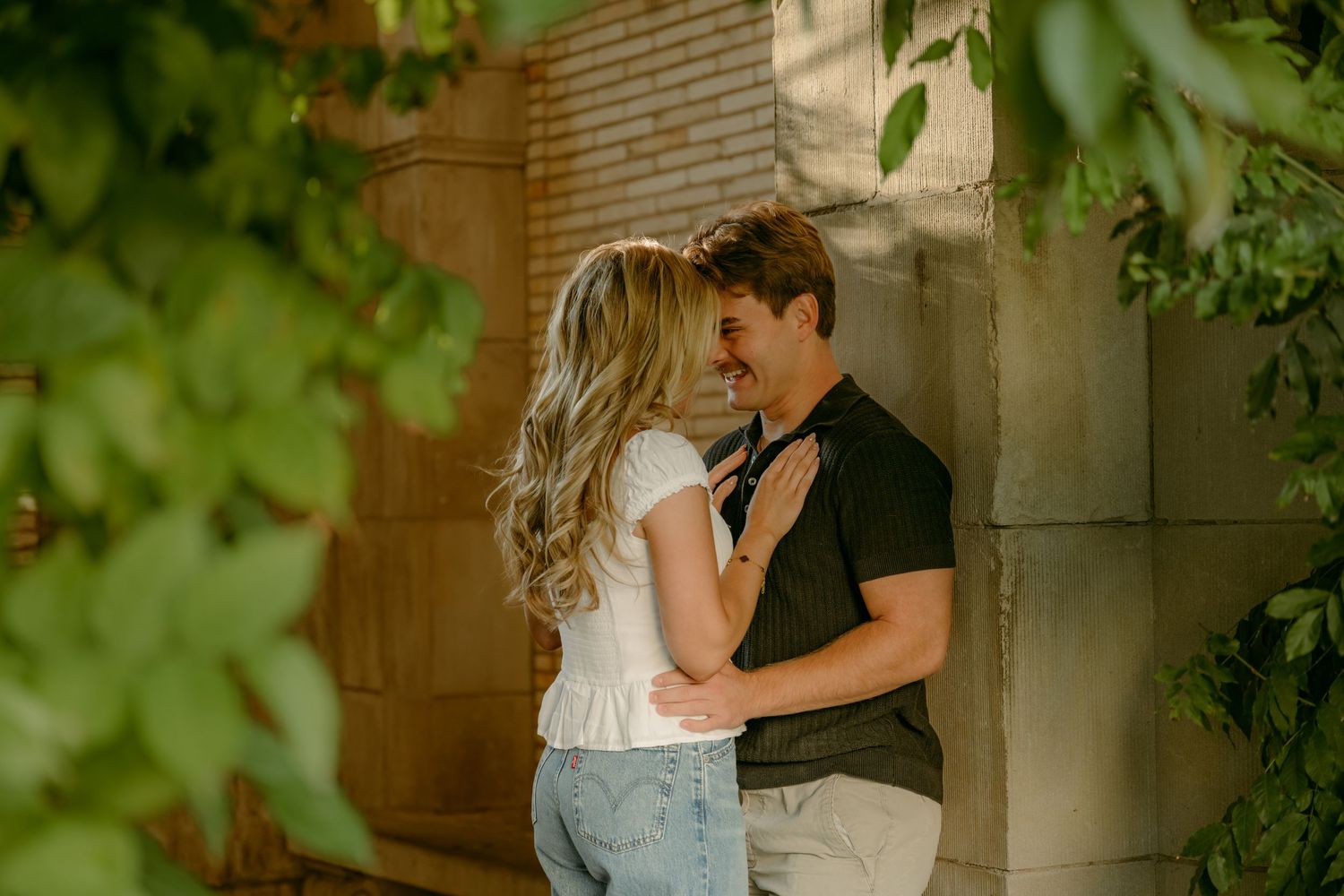 Couple sharing a romantic moment leaning against a stone wall surrounded by lush green foliage.