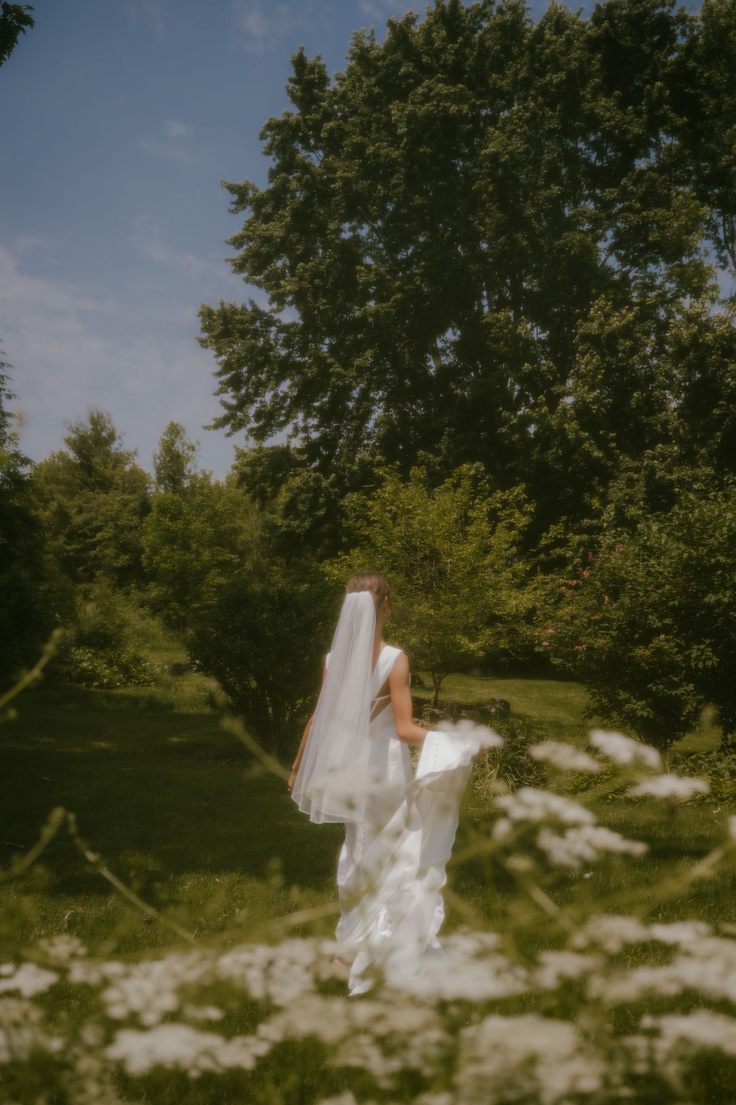 Bride in white gown and veil standing among wildflowers in a sunlit garden on her wedding day.