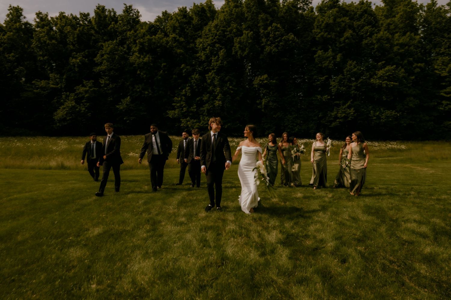 Wedding party walking together across a sunlit green field with dense trees in the background.
