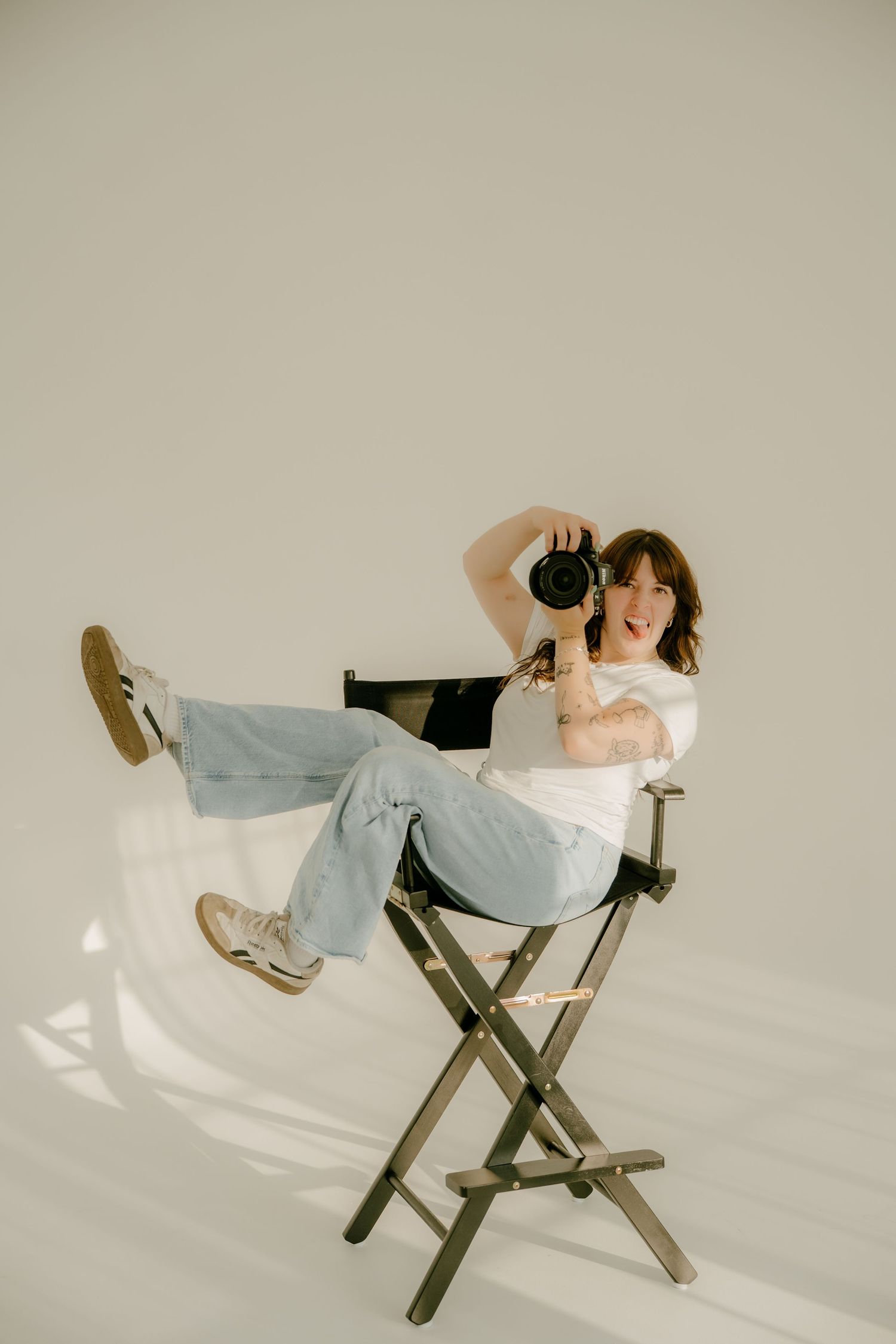 Female photographer sitting casually in director's chair holding camera in bright white studio space.