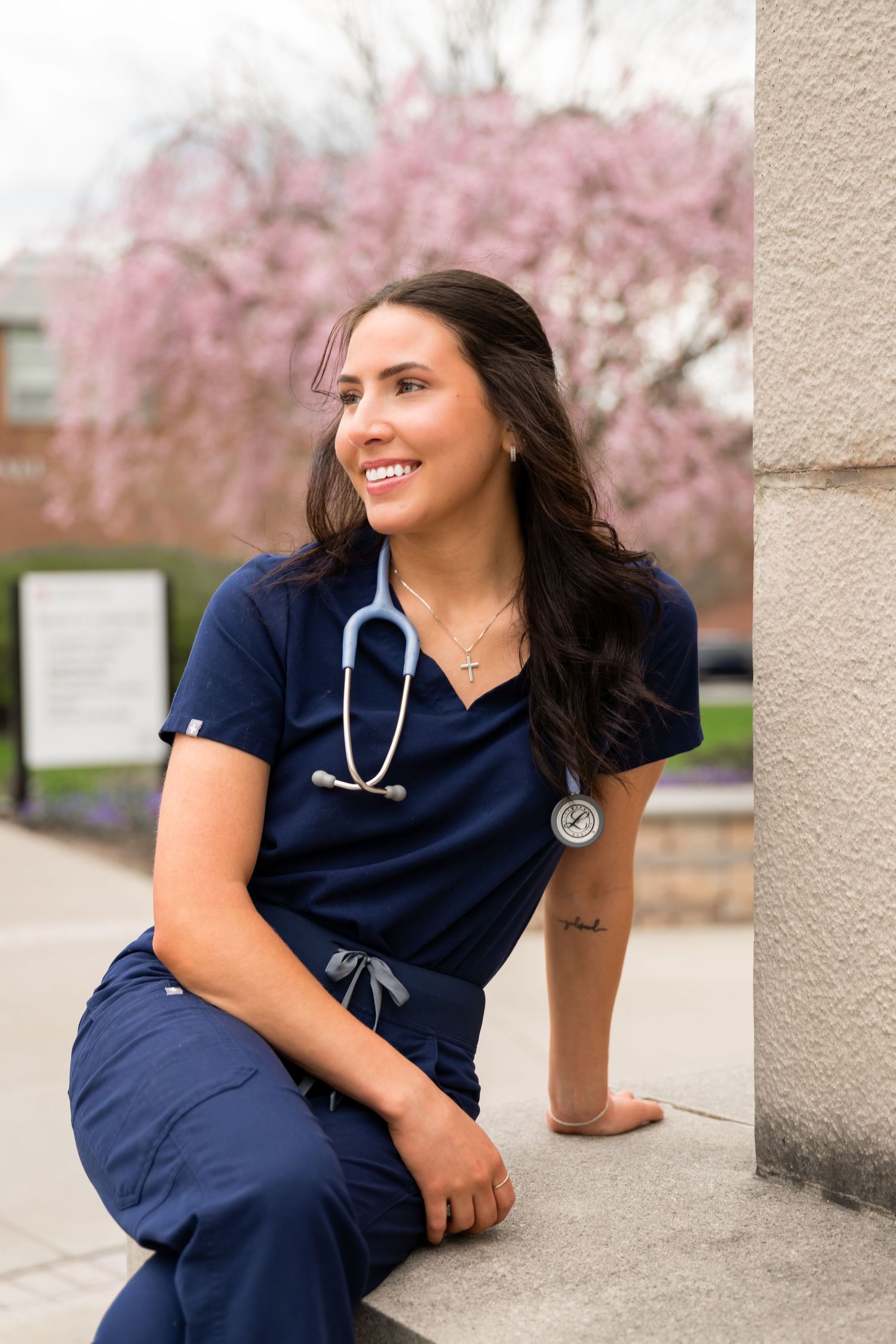 Smiling nurse in navy scrubs with stethoscope leaning against pillar near pink cherry blossom tree.