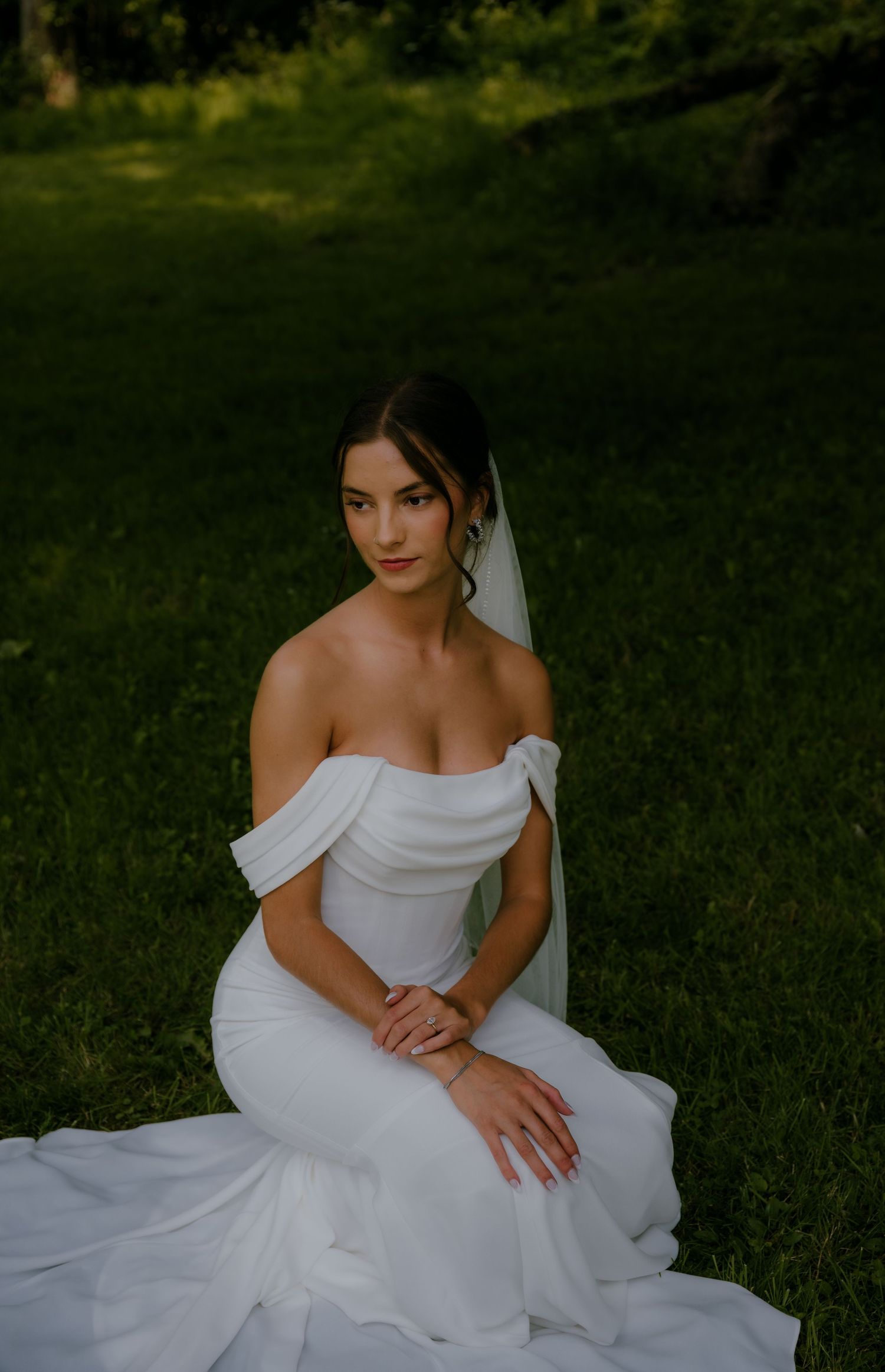 Elegant bride in off-shoulder white gown with veil sitting on green grass in outdoor setting.