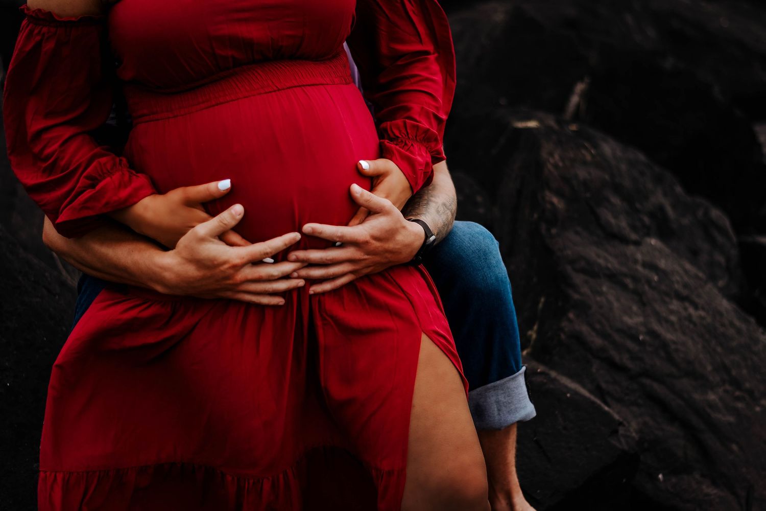 Pregnant woman in red dress with hands cradling baby bump outdoors.