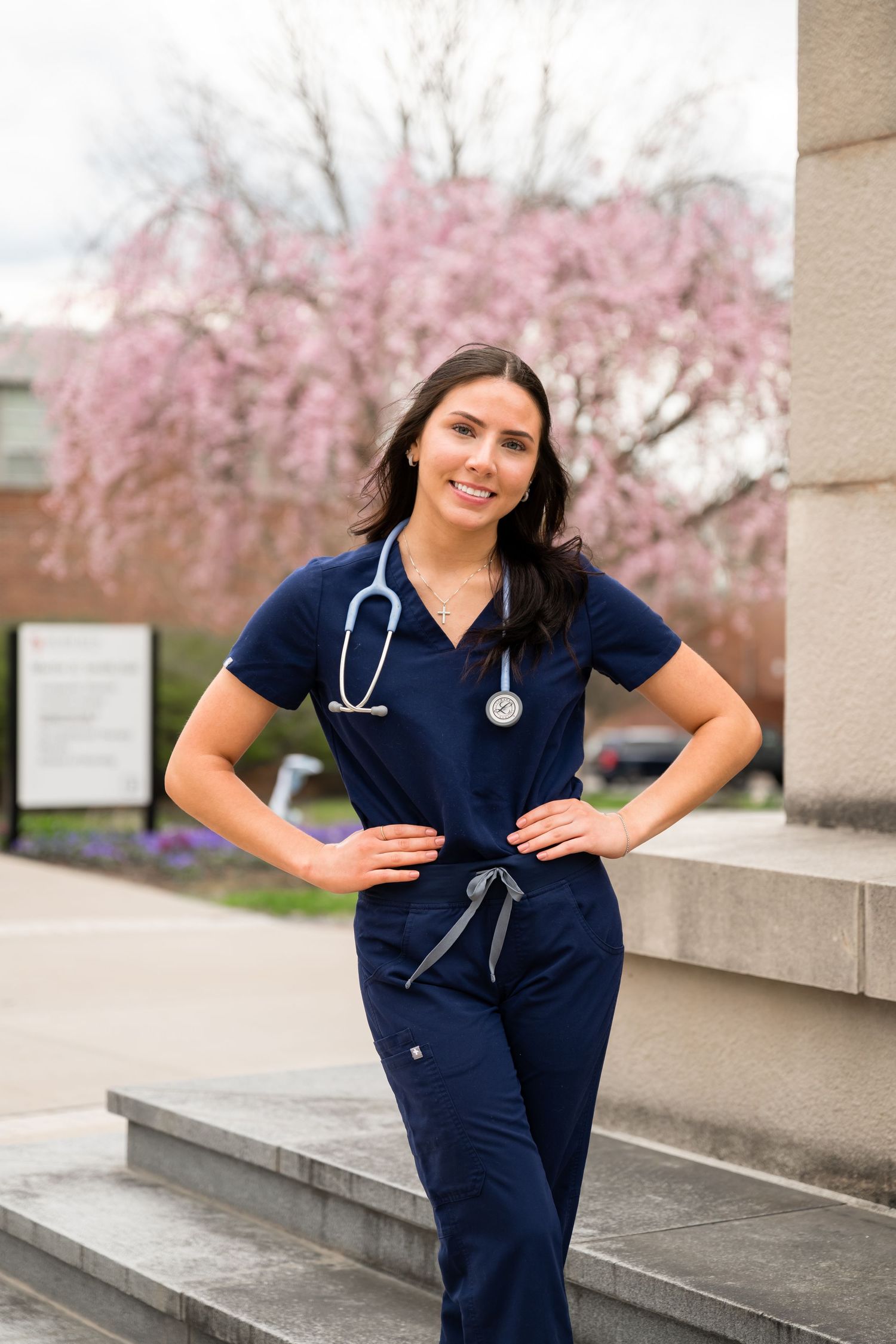 Smiling female nurse in navy scrubs with stethoscope posing outdoors near pink flowering tree.