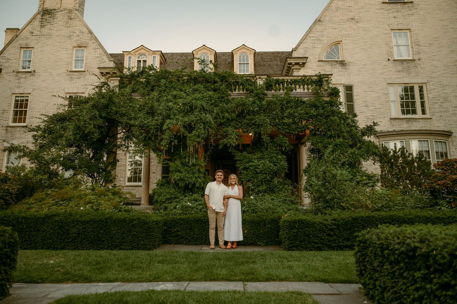 Couple embracing in front of ivy-covered stone mansion at dusk.