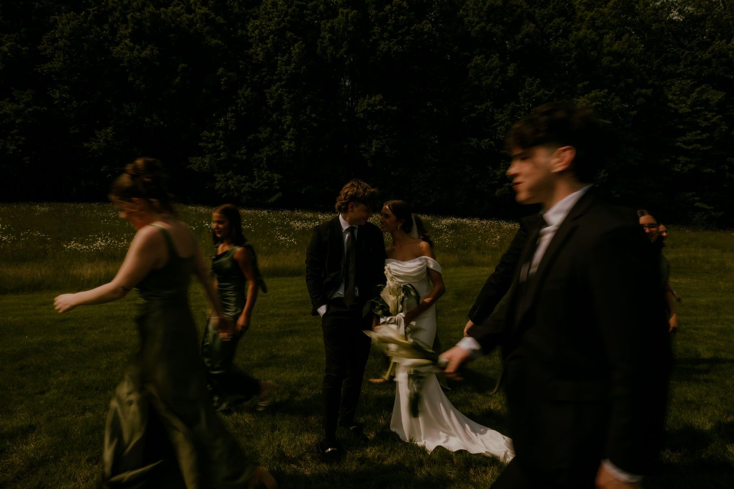 Bride and groom walking with bridesmaids in olive green dresses across a lush green field.