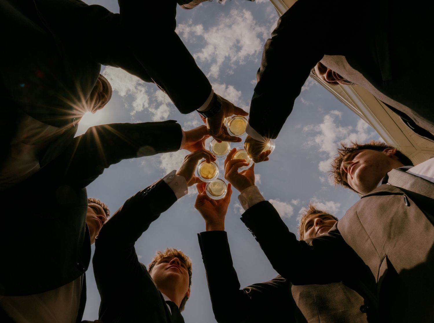 Low angle view of groomsmen toasting drinks in a circle against a bright blue sky.