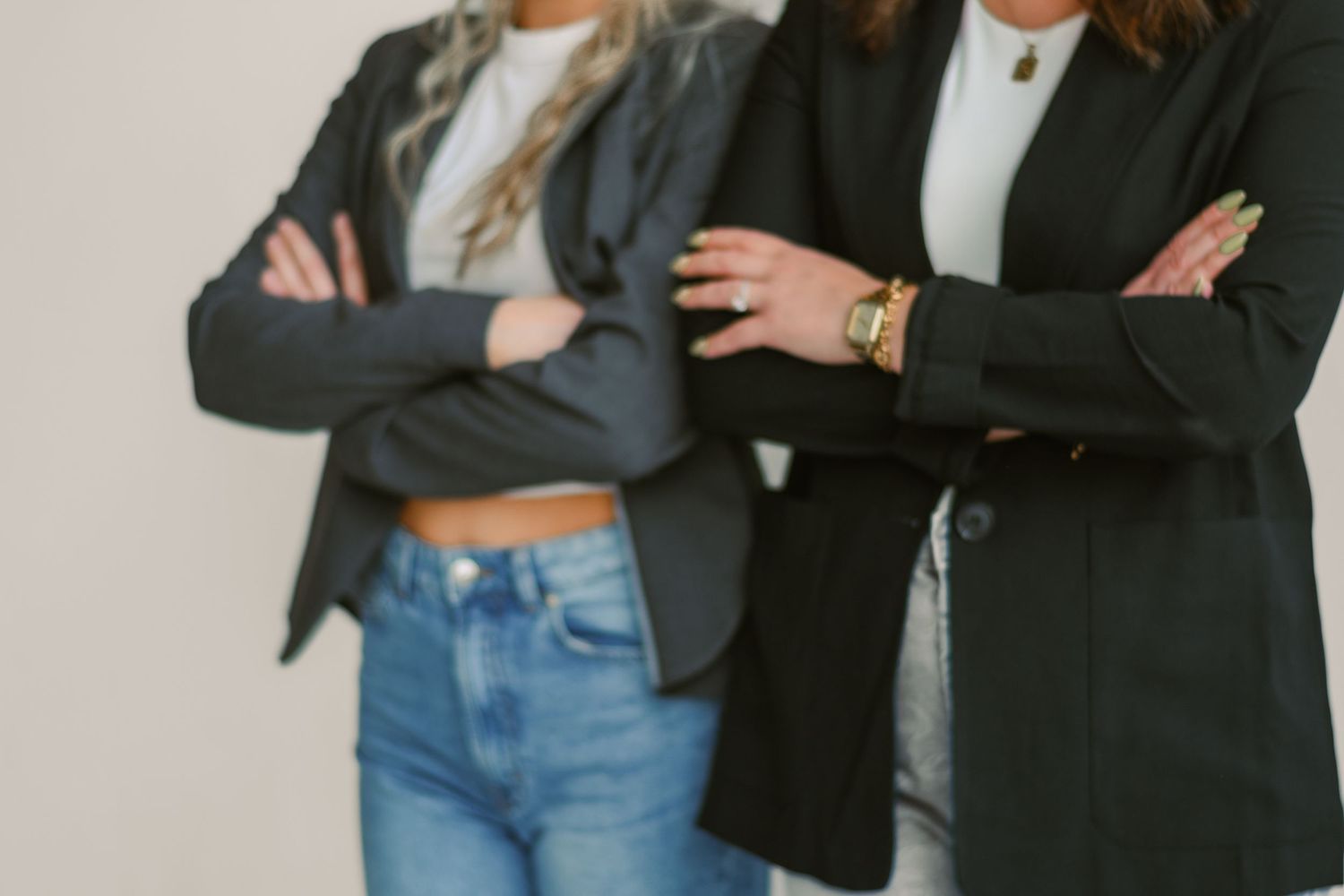 Two women in black blazers standing with arms crossed against a neutral background.