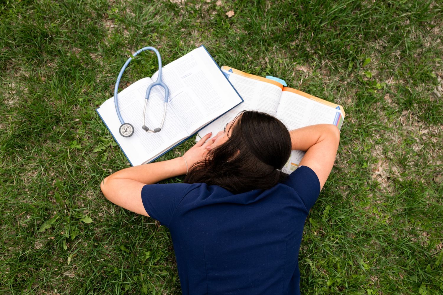 Nursing student resting on grass with stethoscope and open textbook.