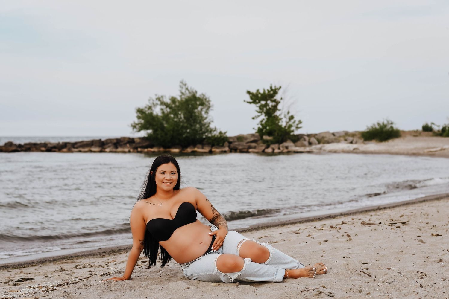 Pregnant woman in black top and white pants sitting relaxed on a sandy beach by the water.
