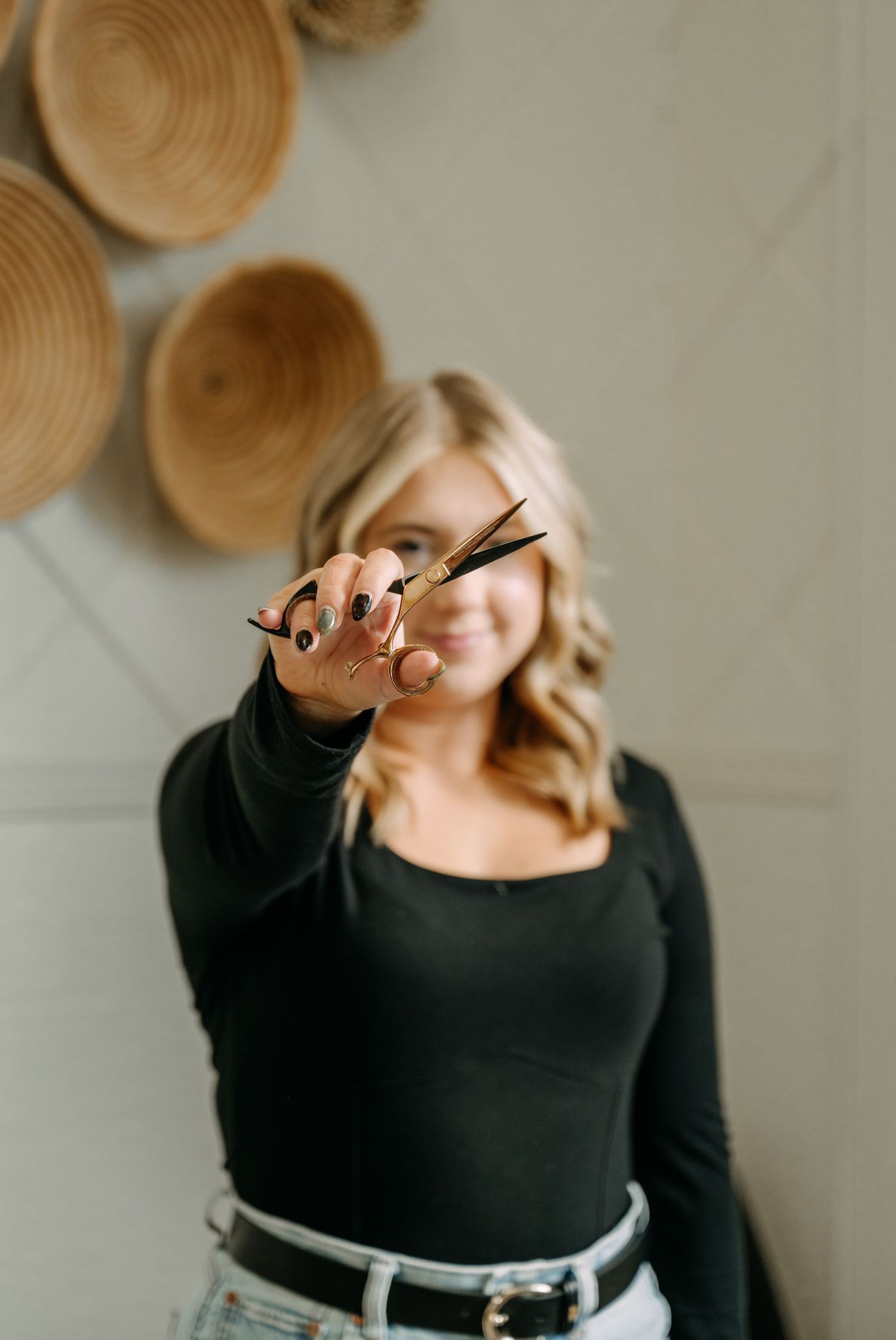 Blonde hairstylist holding gold scissors toward camera in front of woven wall decor.