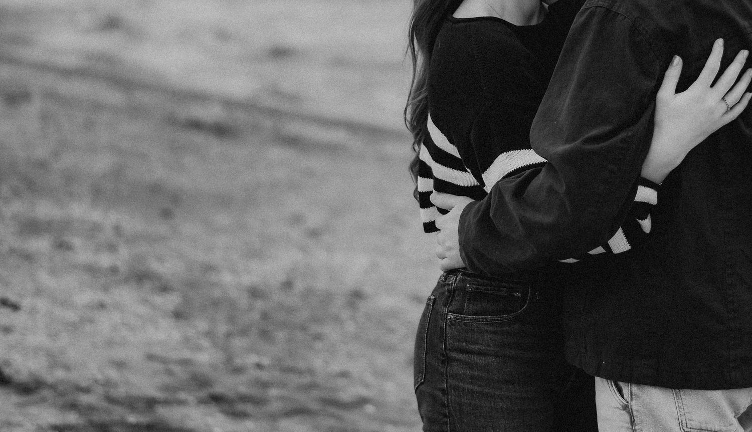 Black and white photo of a couple embracing outdoors, wearing casual striped and denim clothing.
