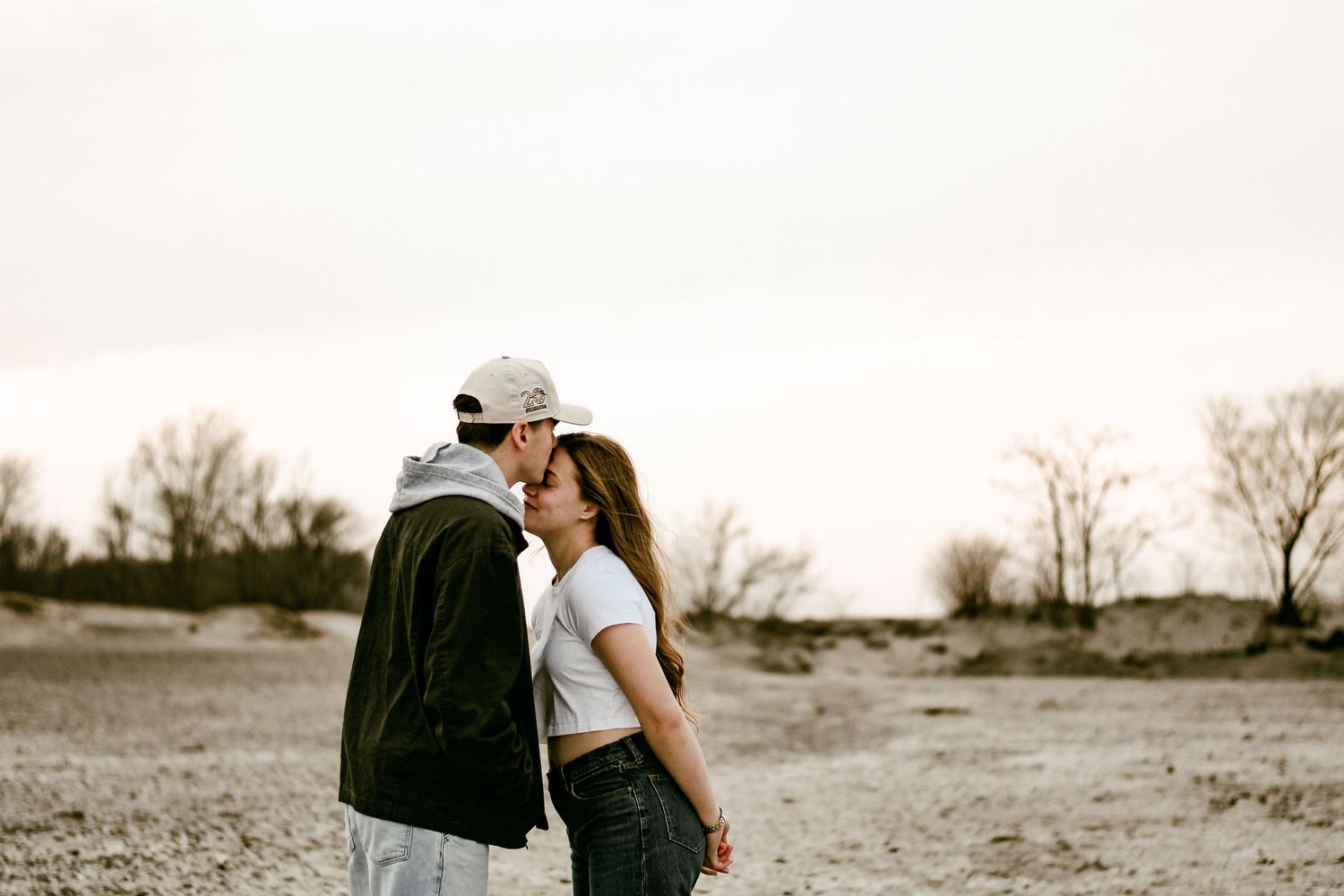 Couple sharing a kiss on a barren landscape at sunset, dressed in casual streetwear.