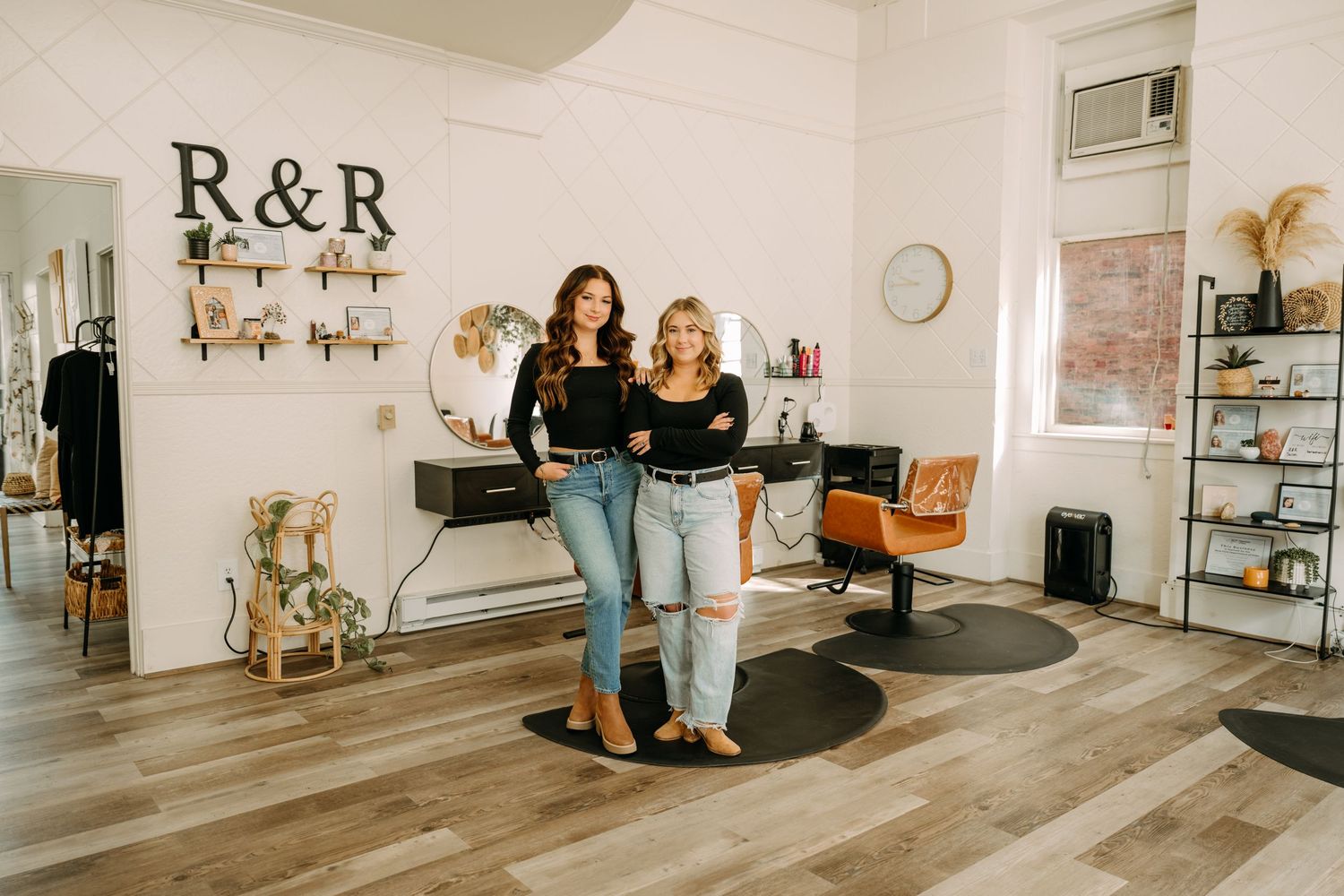 Two female salon owners in black tops and jeans posing in bright modern hair salon.
