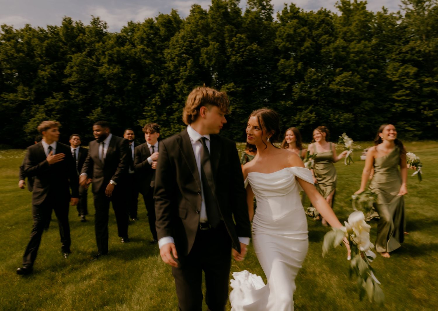 Bride and groom walking hand in hand with wedding party across green lawn outdoors.