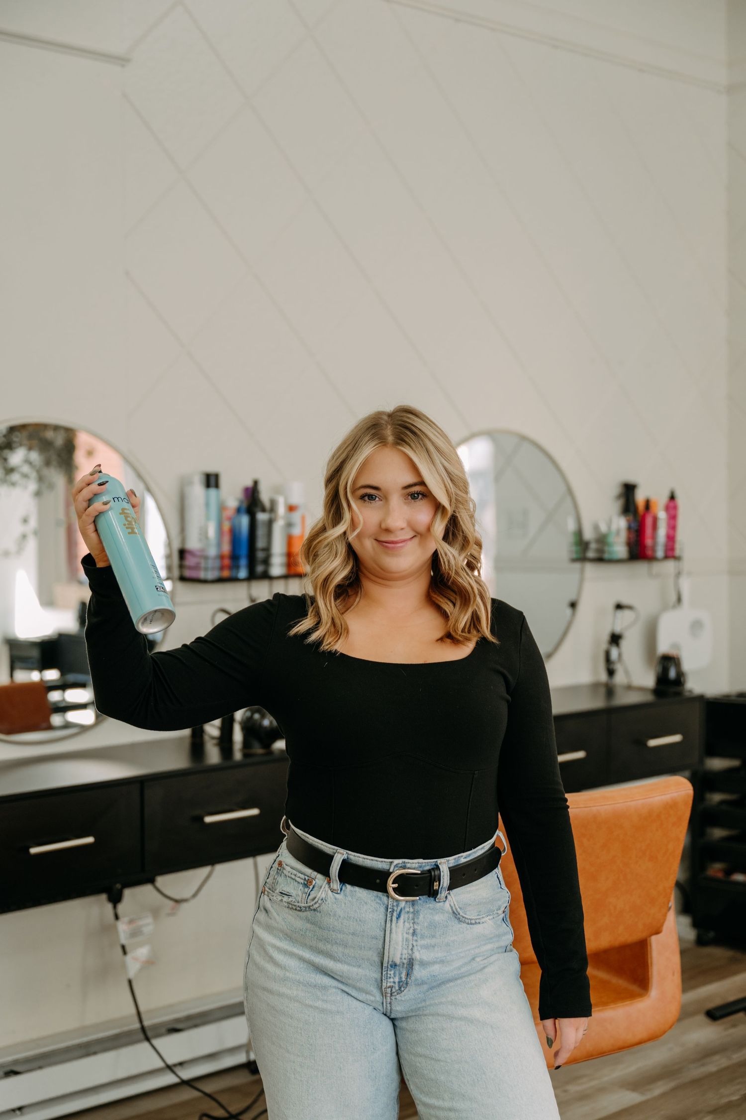 Smiling blonde hairstylist holding hairspray can in modern black and white salon.