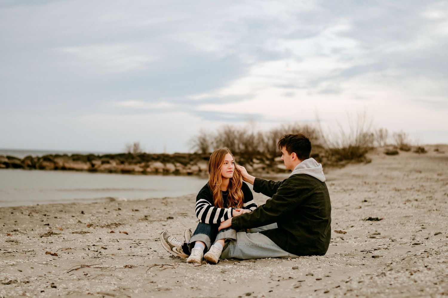 Couple sitting together on a sandy beach at dusk, sharing a tender moment in casual attire.