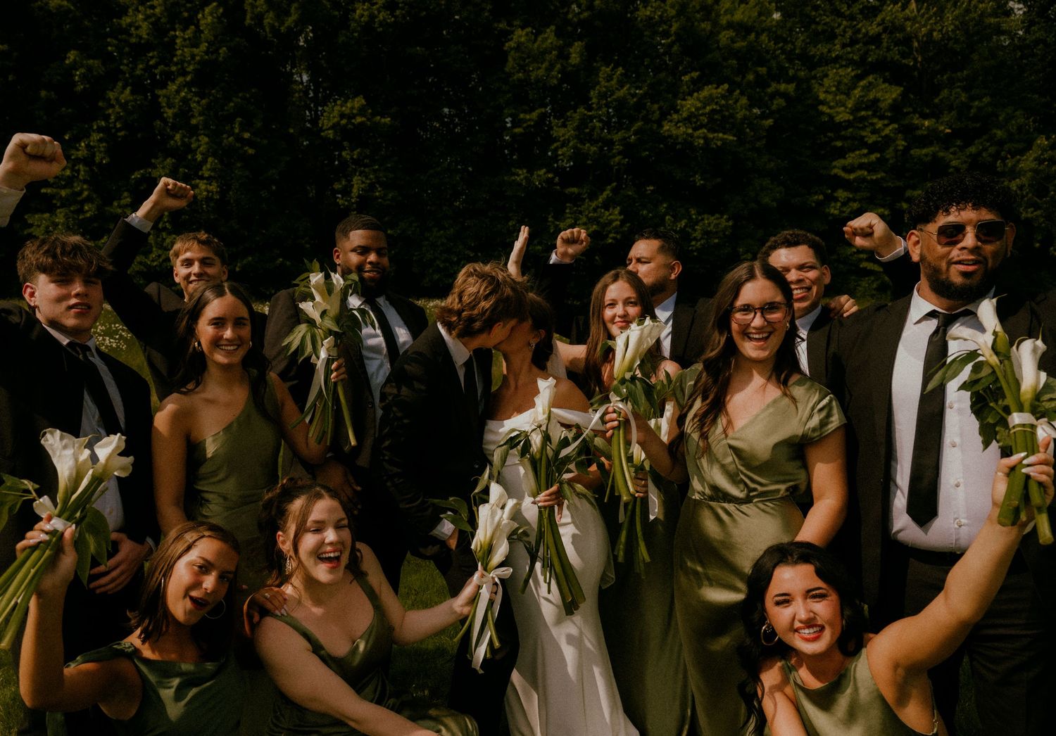 Joyful wedding party celebrating outdoors in green attire with bride holding white floral bouquet.