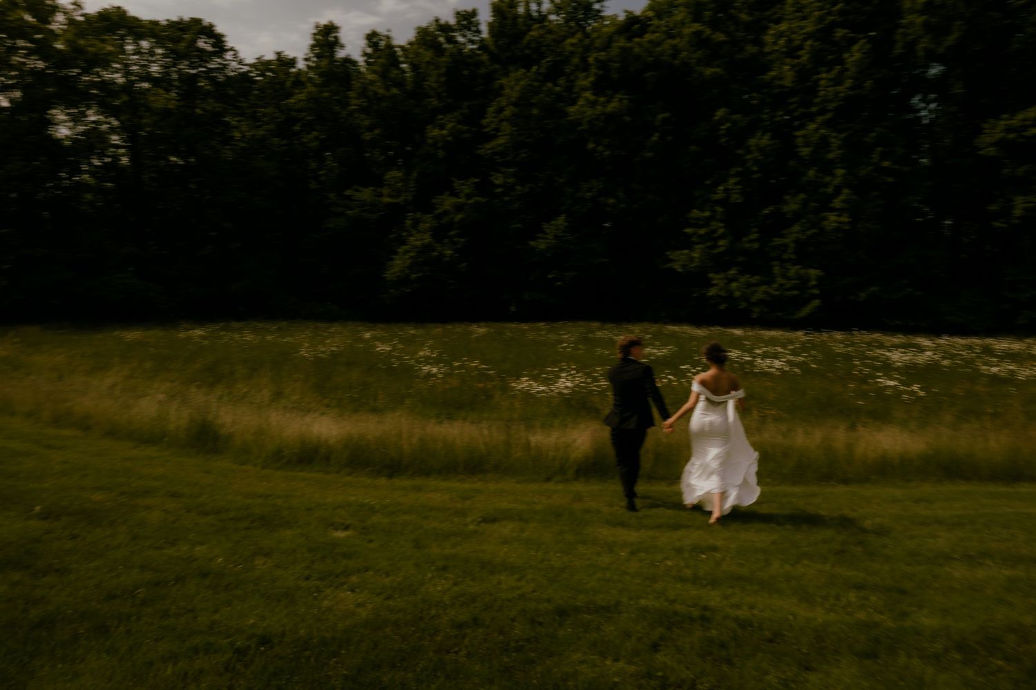 Bride and groom holding hands walking through a green field at golden hour near tall grass.