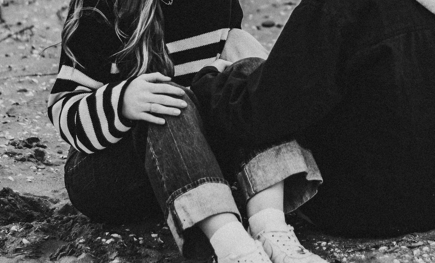Black and white photo of a young woman sitting on the ground in jeans and striped sweater.