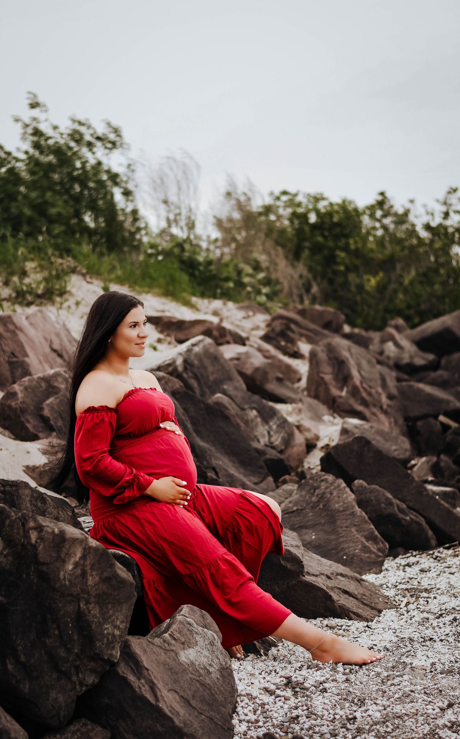 Pregnant woman in a red off-shoulder dress sitting on rocks at a scenic outdoor location.