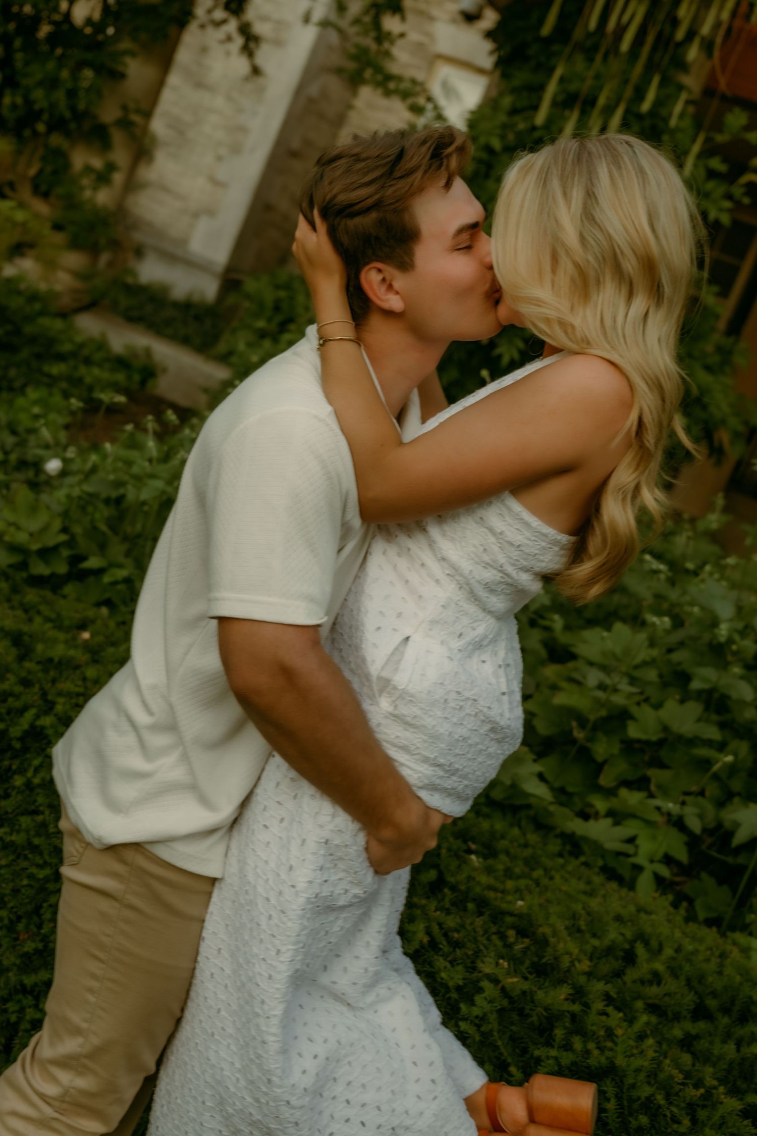 Romantic couple kissing outdoors surrounded by lush green foliage in warm summer light.