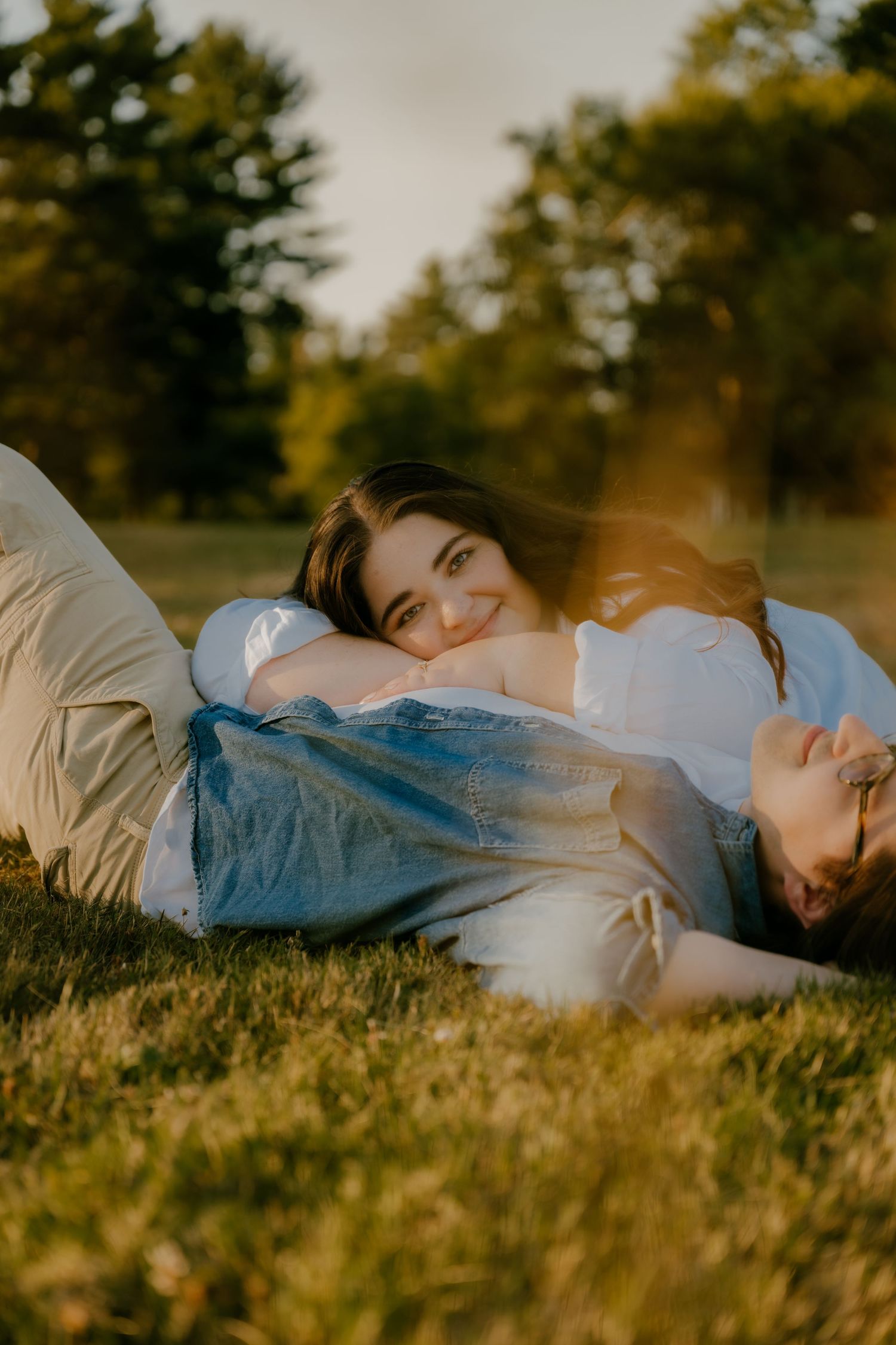 Couple lying on grass in golden sunlight, woman smiling at camera in park setting.