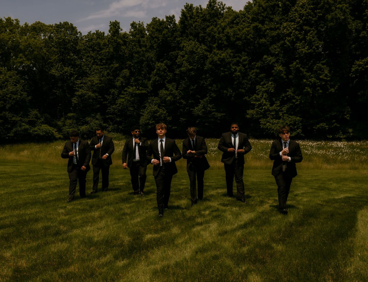 Seven groomsmen in black suits walking across a green field with trees in the background at a wedding.